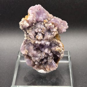 May include: A close-up of a mineral specimen featuring a cluster of light purple crystals. The crystals have a textured, granular appearance, with hints of brown and white. The specimen is displayed on a clear acrylic stand against a dark background.