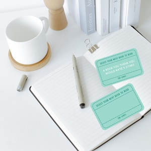 May include: An open notebook with lined pages, a pen, and two teal TBR cards. The cards read "Select Your Next Book to Read" and "A Book You Think You Would Rate 5 Stars". A white mug and books are in the background.