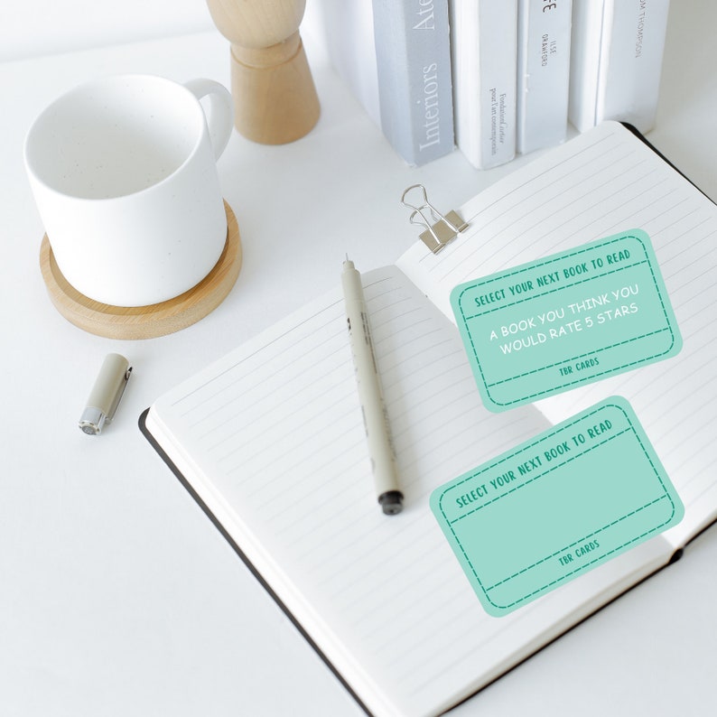 May include: An open notebook with lined pages, a pen, and two teal TBR cards. The cards read "Select Your Next Book to Read" and "A Book You Think You Would Rate 5 Stars". A white mug and books are in the background.