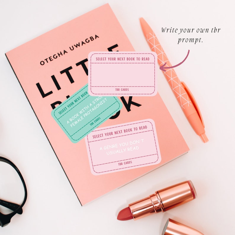 May include: A pink book with the title "LITTLE" and the author's name, OTEGHA UWAGBA. Three TBR cards are on the book, with prompts like "Select your next book to read." A pink pen and lipstick are also visible.