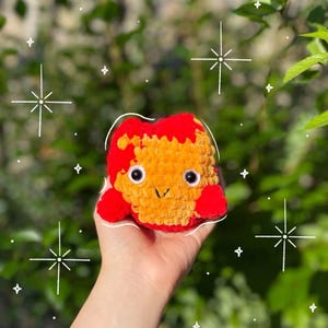 Könnte beinhalten: Ein handgefertigtes, gehäkeltes Plüschtier in Rot- und Orangetönen, das einem Cartoon-Charakter mit großen, runden Augen und einem lächelnden Gesicht ähnelt. Das Plüschtier hat ein strukturiertes, weiches Aussehen und wird in einer Hand vor einem verschwommenen grünen Hintergrund gehalten.