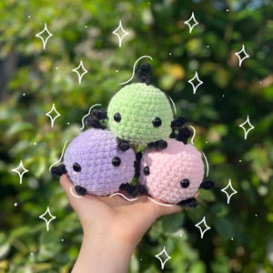 Peut inclure: Trois peluches crochetées aux couleurs pastel : vert clair, lavande et rose. Chacune a des yeux noirs et de petits appendices noirs. Les jouets sont tenus dans une main sur un fond vert flou avec des accents d'étoiles blanches.