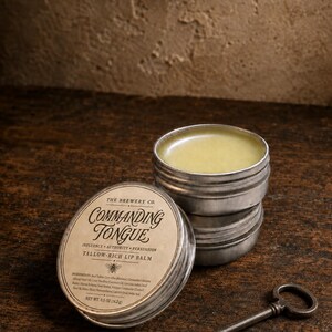 May include: A round metal tin of Commanding Tongue tallow-rich lip balm by The Brewery Co. with a beige label featuring a bee icon and text. The tin is open, revealing a pale yellow balm, and sits next to its lid and a vintage-style metal key.