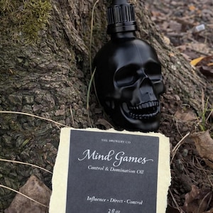 May include: A black skull-shaped bottle with a dropper, next to a label that reads "Mind Games Control & Domination Oil". The label also states "Influence, Direct, Control" and "2 fl oz". The bottle is set against a tree trunk.