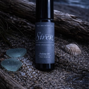 Puede incluir: Un frasco de perfume negro con la palabra "Siren", el texto "THE BREWERY CO" y "Perfume Oil". El frasco está sobre una superficie arenosa con vidrio de mar y una concha. El aceite de perfume es de 10 ml / 0,33 fl oz.