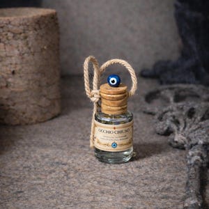 Peut inclure: Petite bouteille en verre avec un bouchon en liège et un charme en forme d'œil bleu. La bouteille porte une étiquette avec du texte et est attachée avec une corde naturelle. Fond gris texturé.