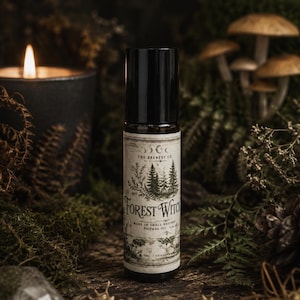 May include: A roll-on perfume oil bottle with a black cap and a label featuring the text "Forest Witch." The label includes illustrations of trees, a crescent moon, and a moth. The bottle is surrounded by moss, ferns, and a lit candle.