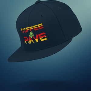 Op de afbeelding: Een marineblauwe baseballcap met de geborduurde tekst "COFFEE & RAVE" in rood, geel en groen. De cap heeft een platte klep en een knoop bovenop. Het ontwerp combineert koffie en rave-cultuur.