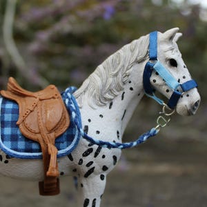 Peut inclure: Un cheval jouet blanc avec des taches noires, une selle marron et un tapis de selle à carreaux bleus. Le cheval a un licou bleu et bleu clair et une longe tressée bleue. Réplique miniature.