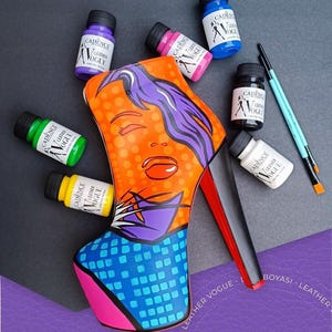 May include: A high-heeled shoe painted with a pop art design of a woman's face. The shoe is surrounded by small bottles of paint in various colours, including yellow, green, blue, pink, black, and white. The shoe is on a purple surface.