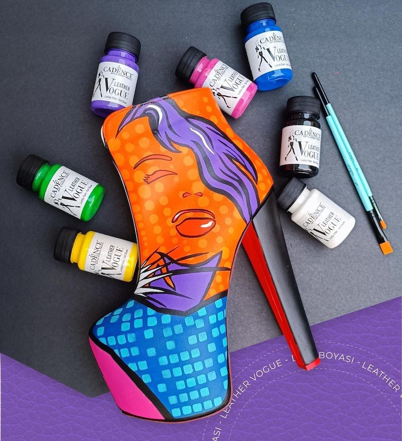 May include: A high-heeled shoe painted with a pop art design of a woman's face. The shoe is surrounded by small bottles of paint in various colours, including yellow, green, blue, pink, black, and white. The shoe is on a purple surface.