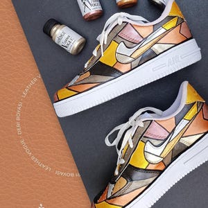 May include: A pair of white trainers with a geometric design painted on them. The design features shades of gold, brown, and silver. The trainers are labelled "AIR" on the side. Four bottles of paint are visible in the background, labelled "CADENCE LEATHER VOGUE".