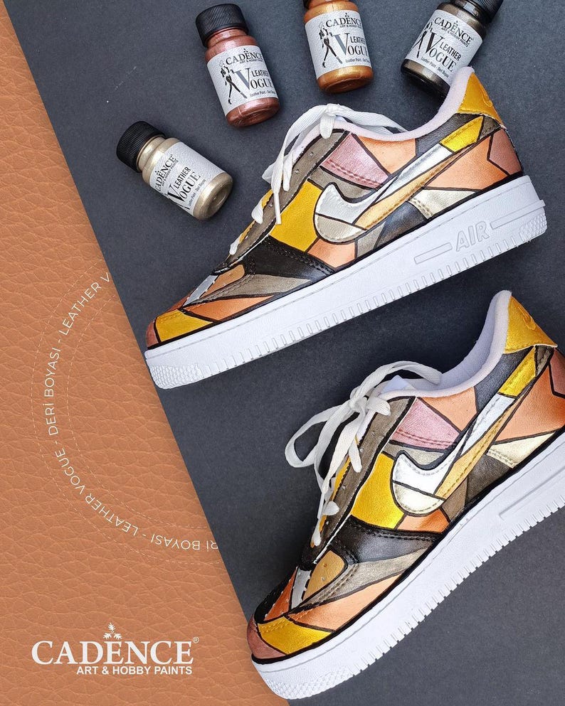 May include: A pair of white trainers with a geometric design painted on them. The design features shades of gold, brown, and silver. The trainers are labelled "AIR" on the side. Four bottles of paint are visible in the background, labelled "CADENCE LEATHER VOGUE".