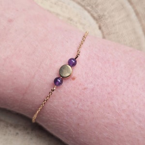 Peut inclure: Un bracelet en chaîne dorée avec trois petites perles violettes et un pendentif rond en or.