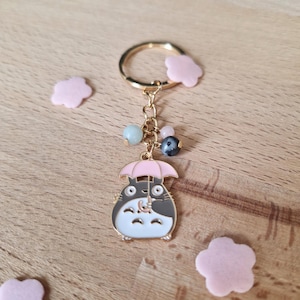 May include: A gold-colored keychain featuring a cartoon character holding a pink umbrella. The character is white with black accents and has large, round eyes. The keychain also includes a gold ring and a chain with several colorful beads.