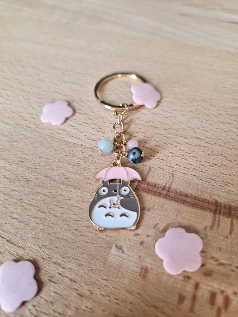 May include: A gold-colored keychain featuring a cartoon character holding a pink umbrella. The character is white with black accents and has large, round eyes. The keychain also includes a gold ring and a chain with several colorful beads.
