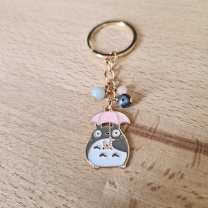 May include: A gold-colored keychain featuring a cartoon character holding a pink umbrella. The keychain has a gold ring and a chain with several colorful beads.