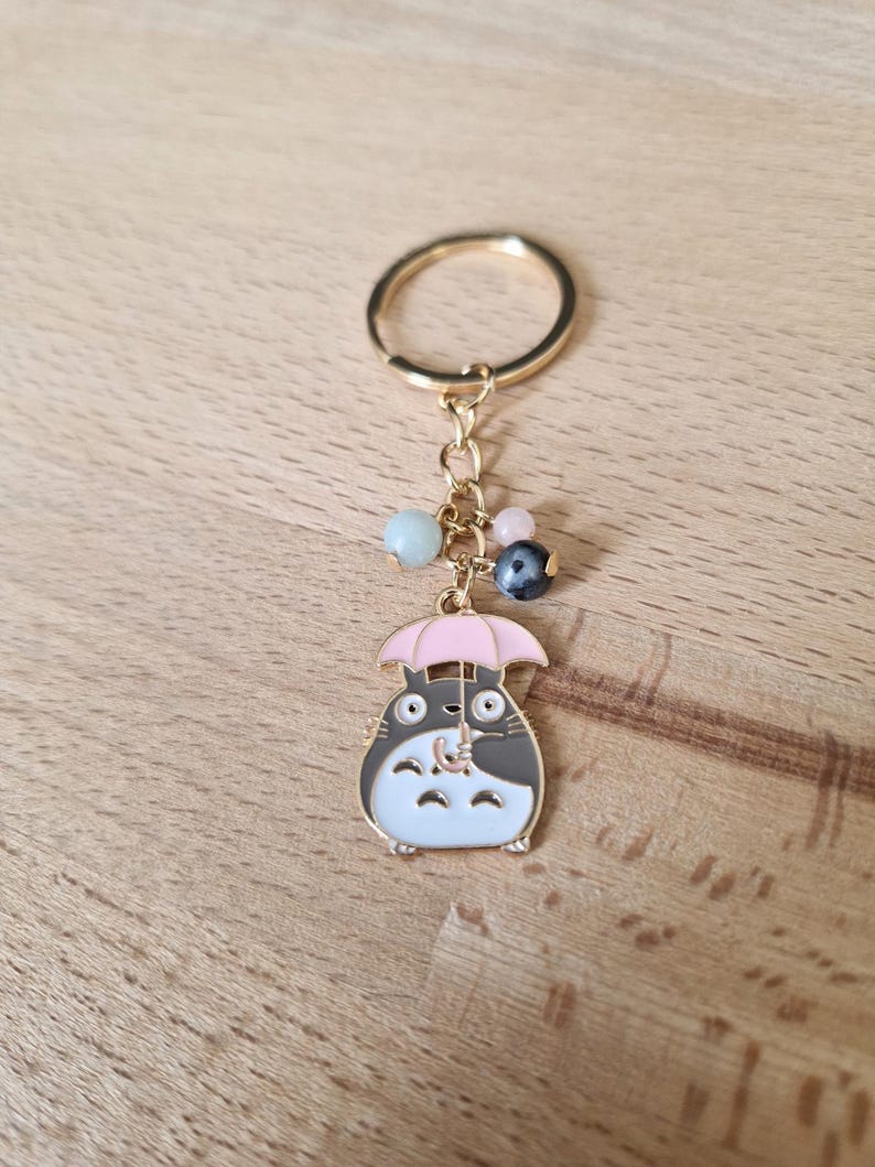 May include: A gold-colored keychain featuring a cartoon character holding a pink umbrella. The keychain has a gold ring and a chain with several colorful beads.