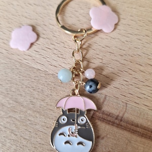 May include: A gold keychain featuring a cartoon character holding a pink umbrella. The character is gray and white with large eyes. The keychain also includes a gold chain with several colorful beads.