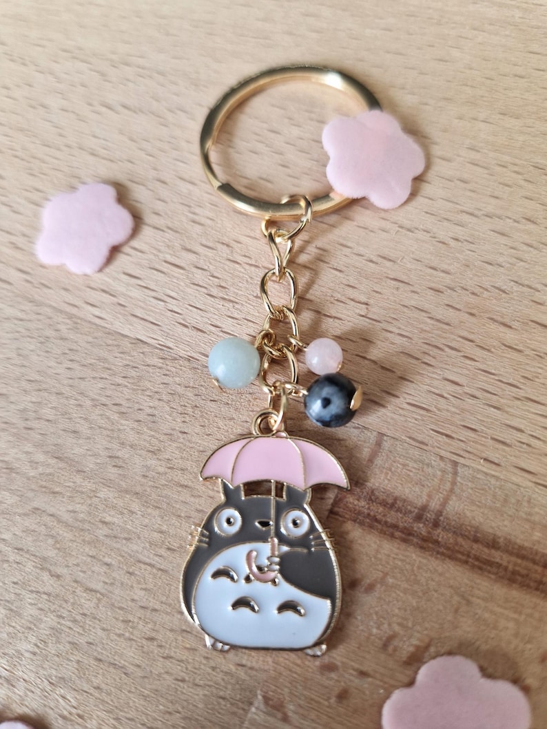May include: A gold keychain featuring a cartoon character holding a pink umbrella. The character is gray and white with large eyes. The keychain also includes a gold chain with several colorful beads.