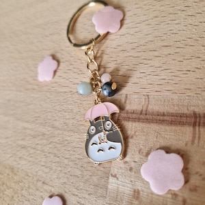 May include: A gold keychain featuring a cartoon character holding a pink umbrella. The character is black and white with a gray belly. The keychain also has a gold ring and several beads in shades of green, pink, and gray.
