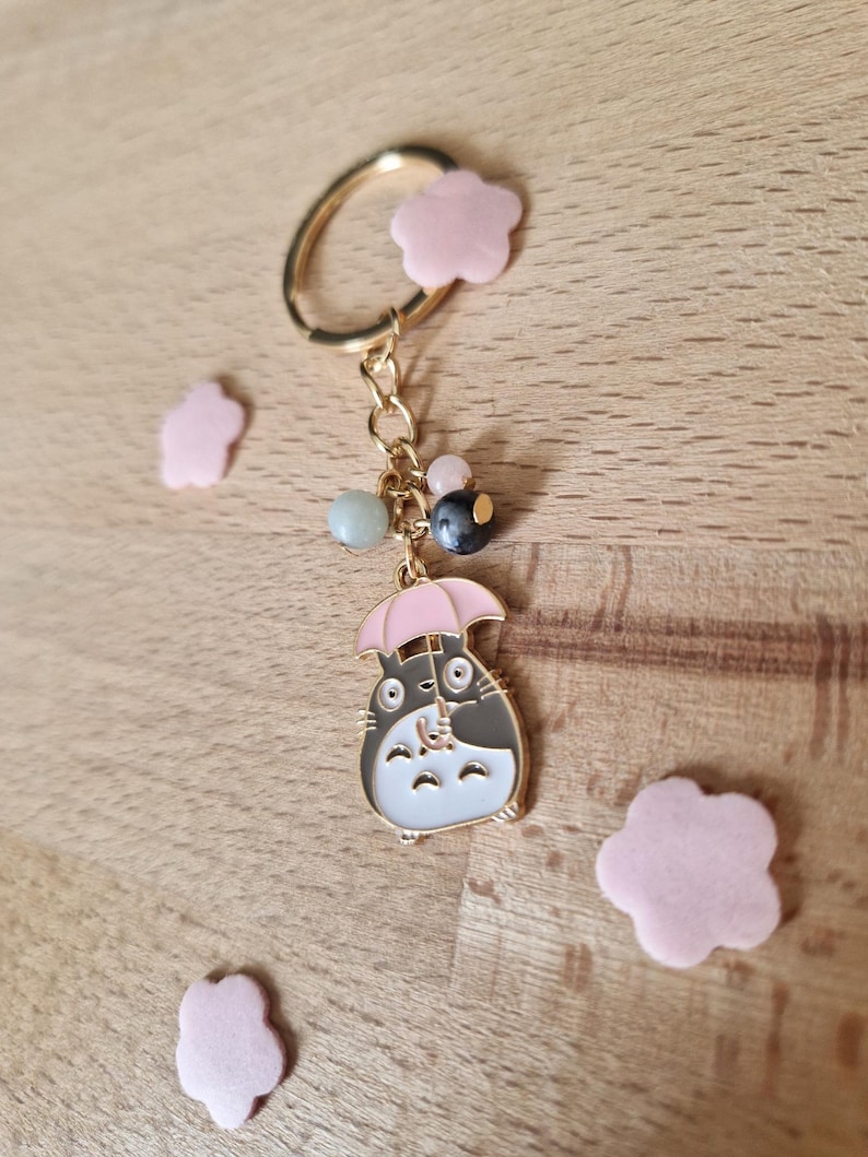 May include: A gold keychain featuring a cartoon character holding a pink umbrella. The character is black and white with a gray belly. The keychain also has a gold ring and several beads in shades of green, pink, and gray.