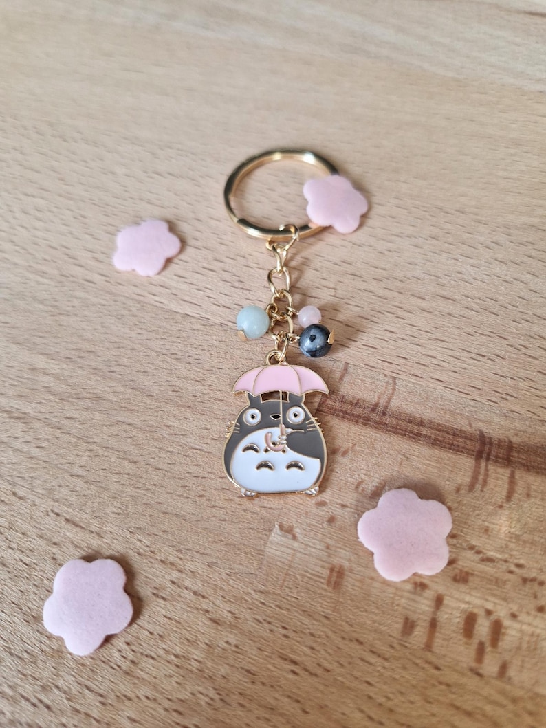May include: A gold-colored keychain featuring a cartoon character holding an umbrella. The character is gray with white accents and has large eyes. The keychain also includes a variety of colorful beads and a pink flower charm.