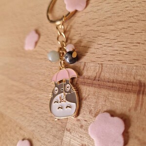 May include: A gold keychain with a grey and white cartoon character holding a pink umbrella. The keychain has a gold chain and several beads, including a green, a black, and a pink bead. The keychain is surrounded by pink flower-shaped charms.