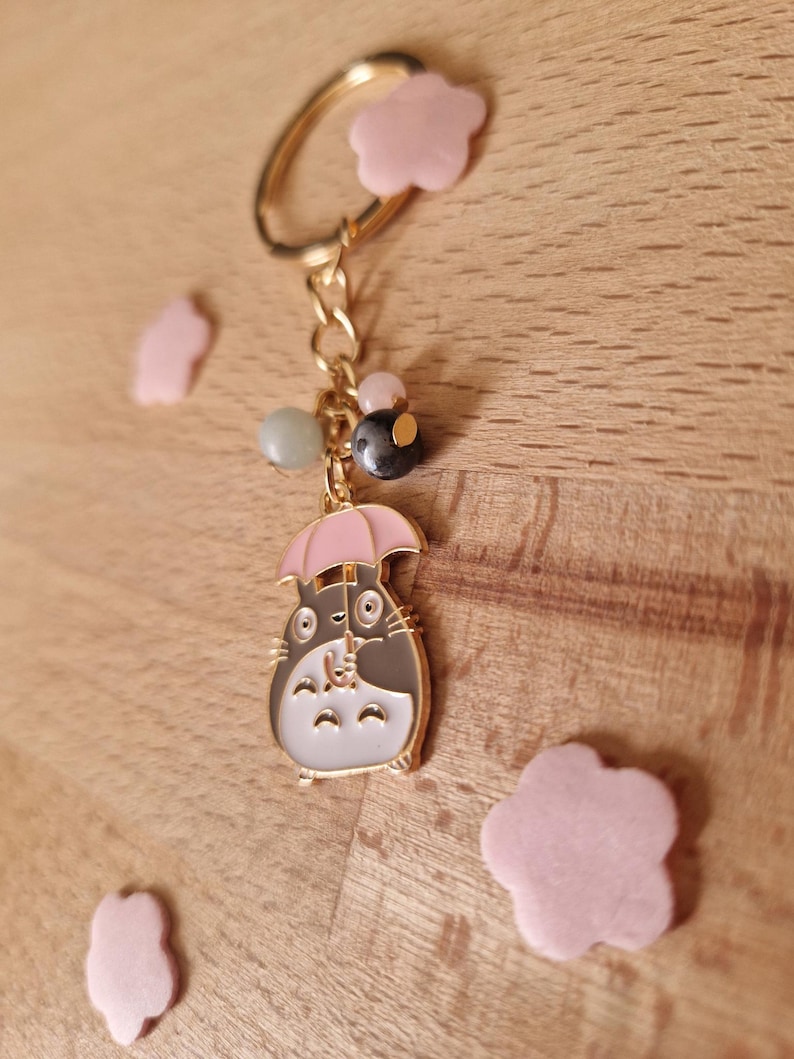 May include: A gold keychain with a grey and white cartoon character holding a pink umbrella. The keychain has a gold chain and several beads, including a green, a black, and a pink bead. The keychain is surrounded by pink flower-shaped charms.