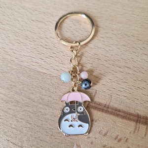 May include: Gold keychain featuring a cartoon character holding a pink umbrella. The character is black and white with a brown nose and has a small, white, fluffy tail. The keychain has a gold ring and a chain with colorful beads.
