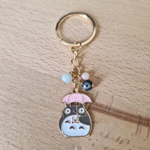 May include: Gold keychain featuring a cartoon character holding a pink umbrella. The character is black and white with a brown nose and has a small, white, fluffy tail. The keychain has a gold ring and a chain with colorful beads.