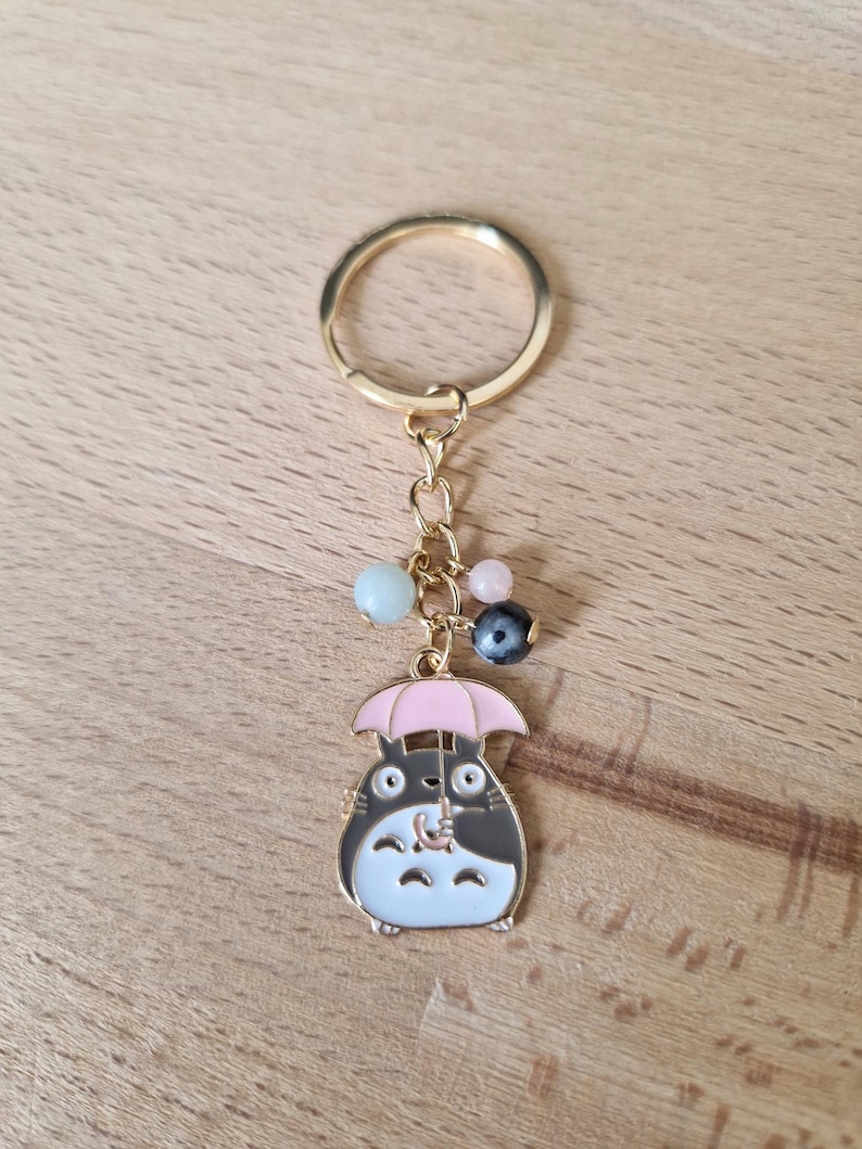 May include: Gold keychain featuring a cartoon character holding a pink umbrella. The character is black and white with a brown nose and has a small, white, fluffy tail. The keychain has a gold ring and a chain with colorful beads.
