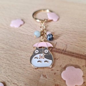 May include: A gold-colored keychain featuring a cartoon character holding a pink umbrella. The character is white with black accents and has large eyes. The keychain also includes a gold ring and several colorful beads.