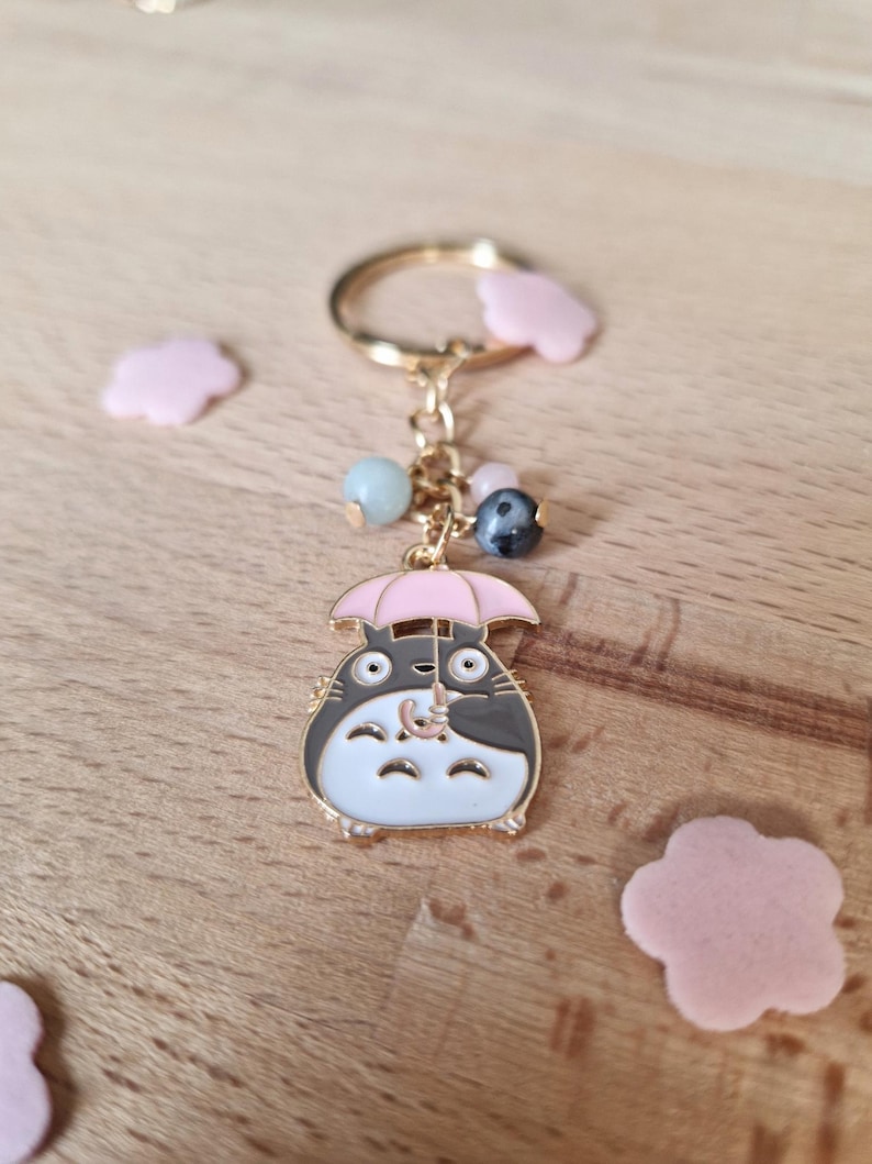 May include: A gold-colored keychain featuring a cartoon character holding a pink umbrella. The character is white with black accents and has large eyes. The keychain also includes a gold ring and several colorful beads.