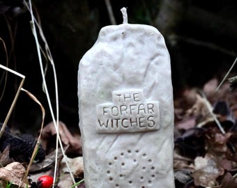 Op de afbeelding: Een witte kaars in de vorm van een grafsteen met de tekst "The Forfar Witches" en "Just People" erop. De kaars staat op een bed van bruine bladeren en rode bessen.