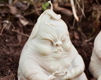 Op de afbeelding: Een witte keramische kaars in de vorm van een mediterend figuur met een vlam erop. Het figuur zit op een bed van bruine aarde.