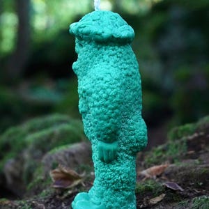 Op de afbeelding: Een tealgroene kaars in de vorm van een persoon met een hoed. De kaars heeft een getextureerd oppervlak en staat op een mossige boomstam.