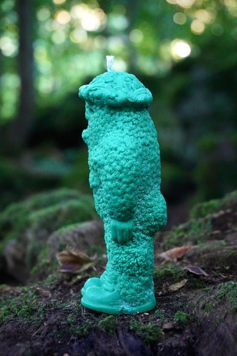 Op de afbeelding: Een tealgroene kaars in de vorm van een persoon met een hoed. De kaars heeft een getextureerd oppervlak en staat op een mossige boomstam.