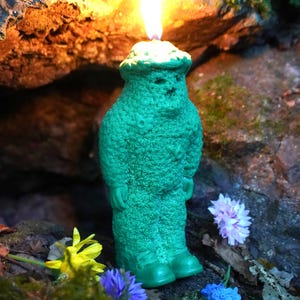 Op de afbeelding: Een groene kaars in de vorm van een persoon met een vlam erop. De kaars is omringd door bloemen en staat op een rots.