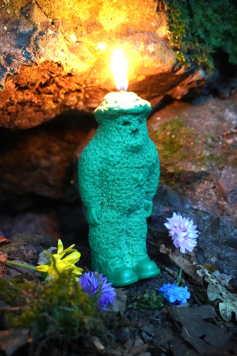 Op de afbeelding: Een groene kaars in de vorm van een persoon met een vlam erop. De kaars is omringd door bloemen en staat op een rots.