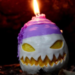 May include: A purple and white Halloween candle shaped like a jack-o'-lantern with a lit flame on top.