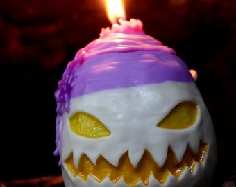 Op de afbeelding: Een paarse en witte Halloween-kaars in de vorm van een jack-o'-lantern met een brandende vlam erop.