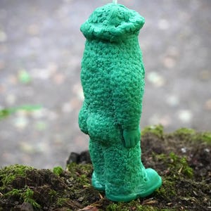 Op de afbeelding: Een groene kaars in de vorm van een persoon met een hoed. De kaars heeft een getextureerd oppervlak en staat op een bed van mos.