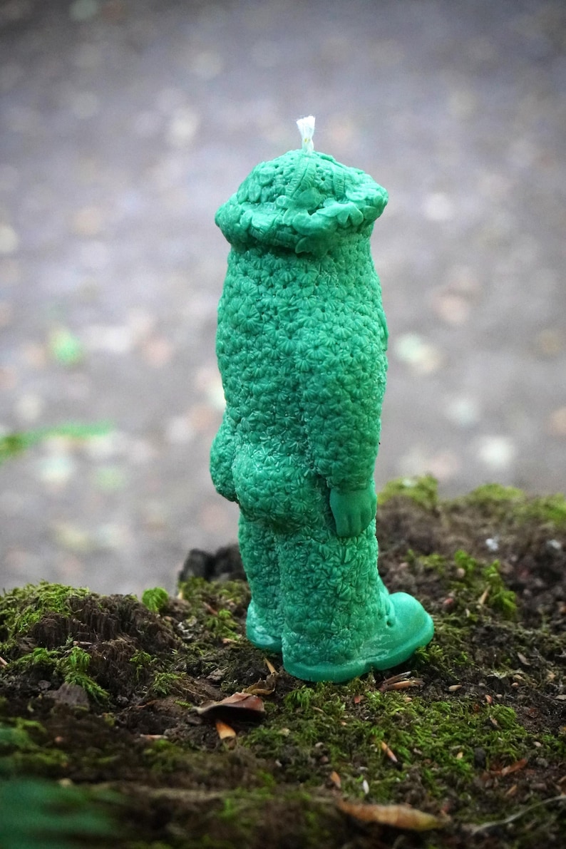 Op de afbeelding: Een groene kaars in de vorm van een persoon met een hoed. De kaars heeft een getextureerd oppervlak en staat op een bed van mos.
