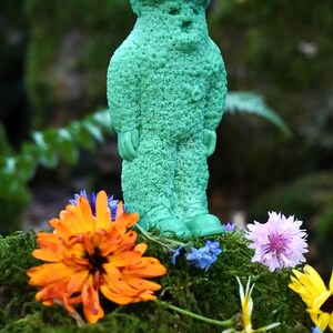 Op de afbeelding: Een groene kaars in de vorm van een persoon met een textuur, staand op een bed van mos met oranje, paarse en gele bloemen.