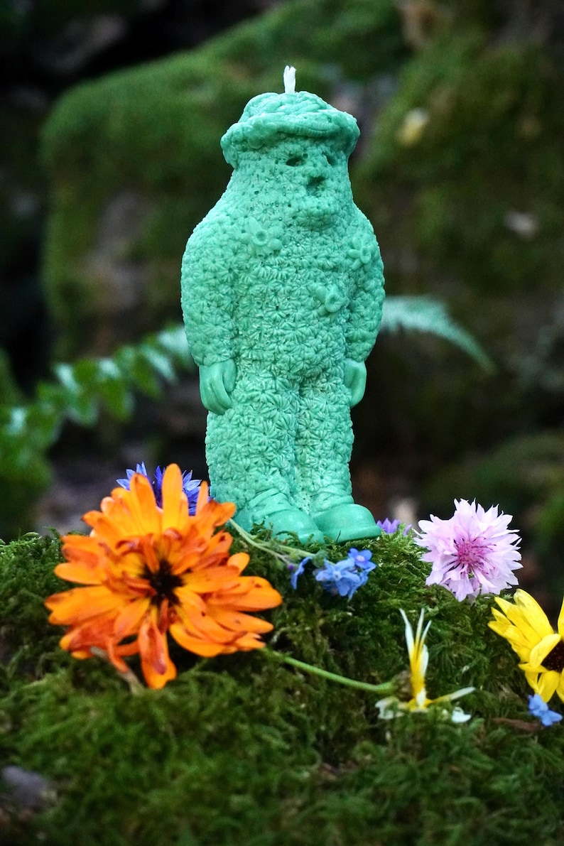Op de afbeelding: Een groene kaars in de vorm van een persoon met een textuur, staand op een bed van mos met oranje, paarse en gele bloemen.