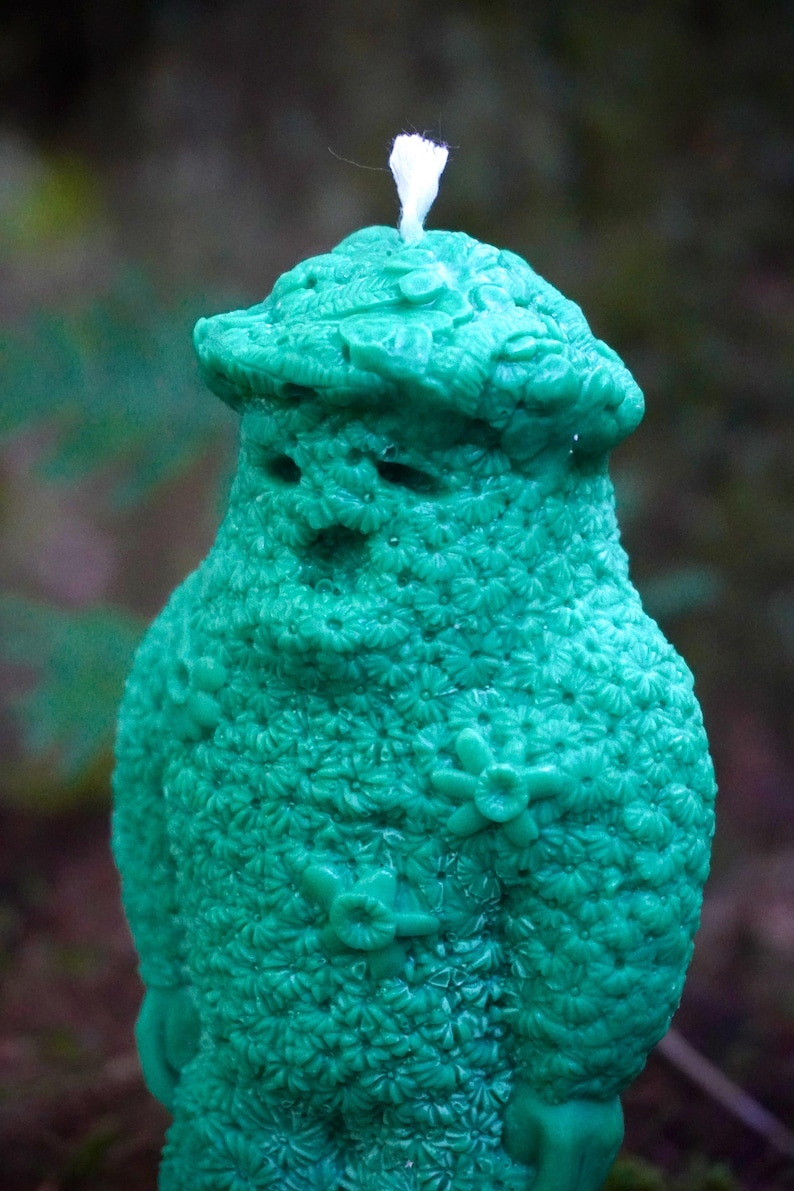 Op de afbeelding: Een groene kaars in de vorm van een persoon met een witte lont. De kaars is bedekt met een bloemenpatroon.