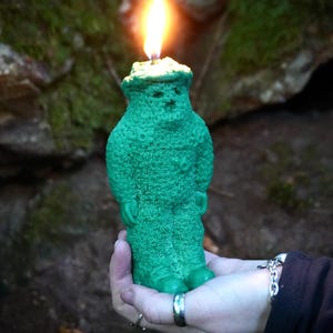 Op de afbeelding: Een groene kaars in de vorm van een persoon met een hoed en een vlam erbovenop. De kaars wordt in een hand gehouden.