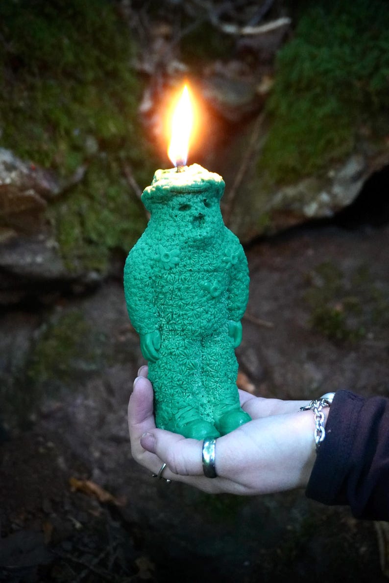 Op de afbeelding: Een groene kaars in de vorm van een persoon met een hoed en een vlam erbovenop. De kaars wordt in een hand gehouden.
