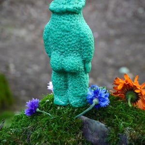 Op de afbeelding: Een groene kaars in de vorm van een persoon met een getextureerd oppervlak. De kaars staat op een bed van mos met een paar bloemen eromheen.
